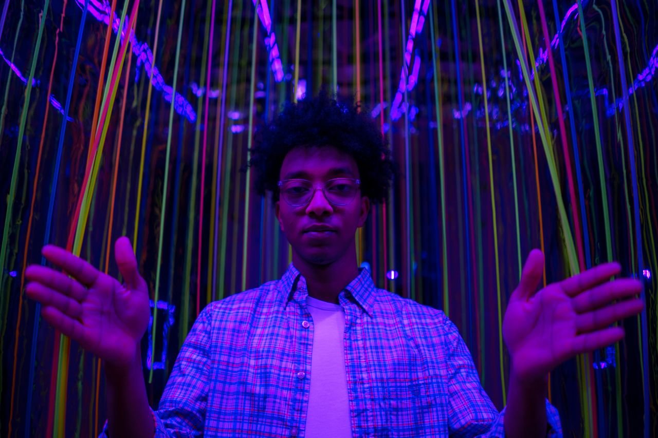 services-04 Man with afro hairstyle standing among neon strings in a dark room, creating a futuristic and vibrant atmosphere.